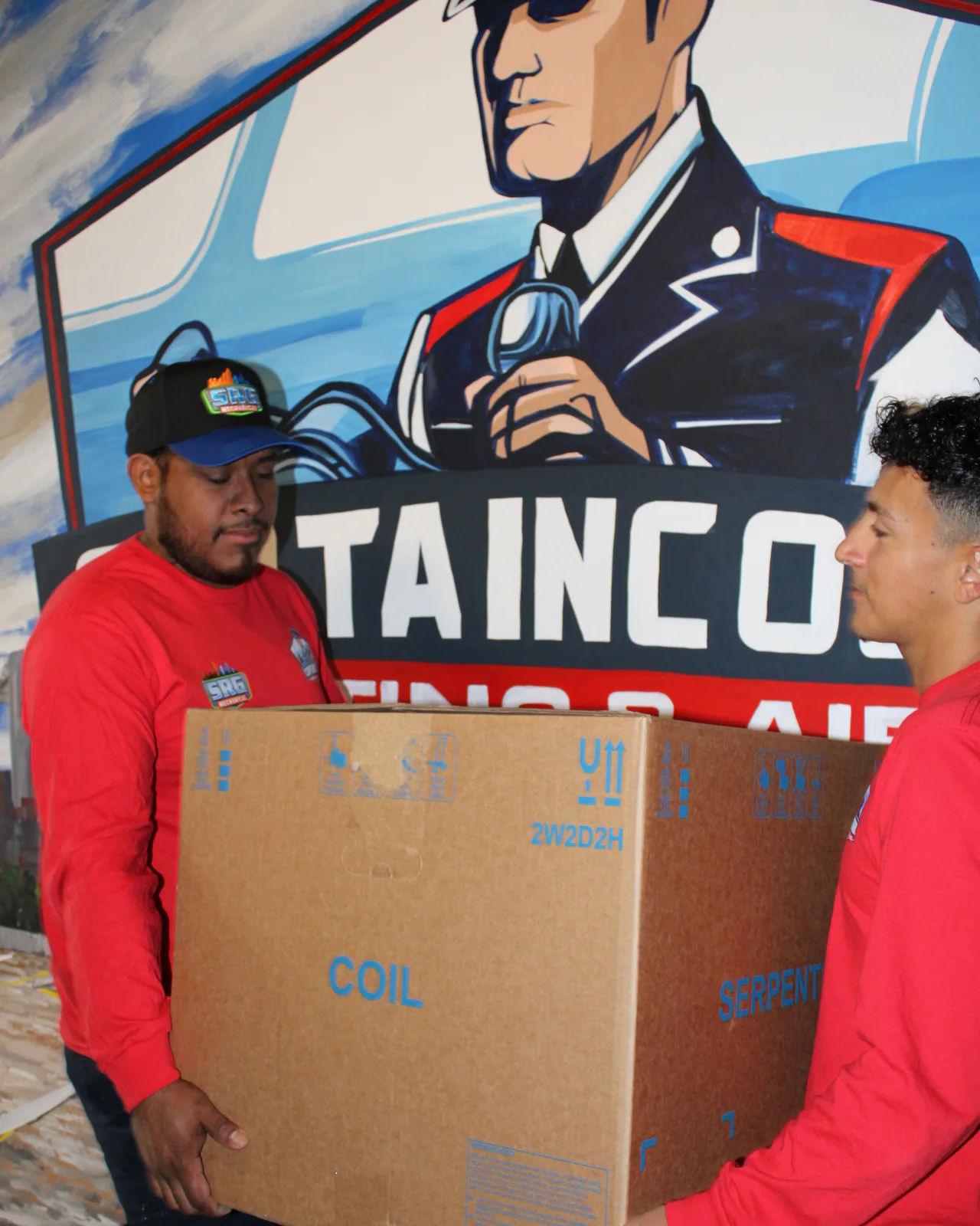 Two Captain Cool Heating & Air technicians carrying a boxed evaporator coil past the captain mural
